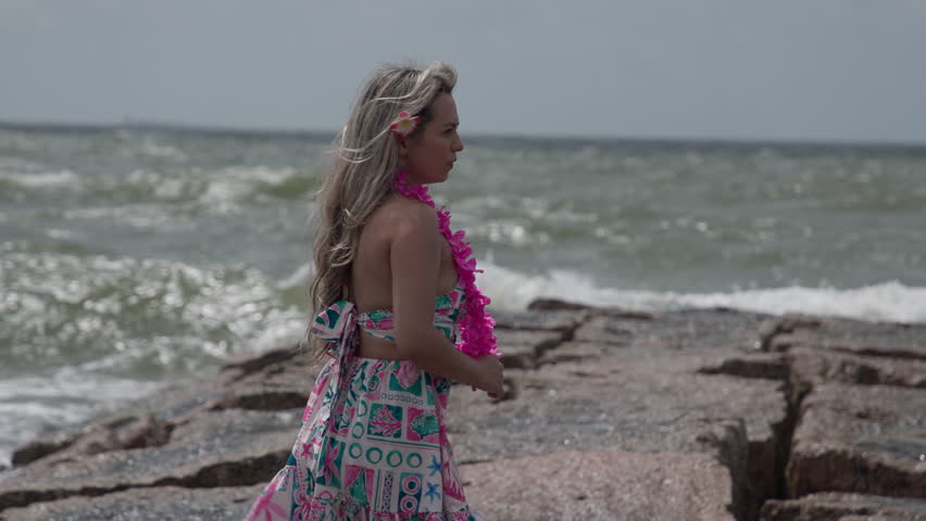 A young woman in a colorful summer dress stands on a rocky pier at Galveston Beach, gazing at crashing ocean waves under a cloudy, windswept sky.