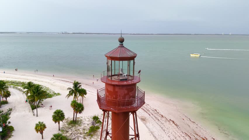 Aerial view of the historic Sanibel Island Lighthouse standing on the sandy beach with palm trees and calm ocean waters in Lee County, Florida, United States.