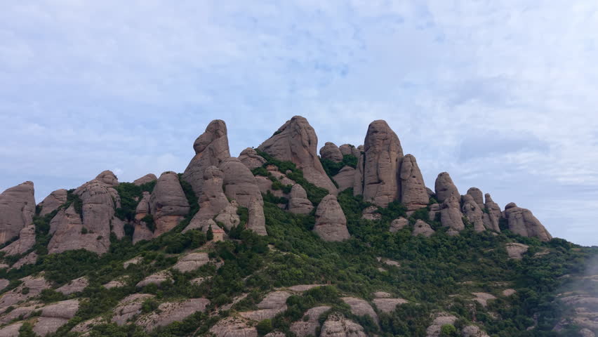 Conglomerate rock peaks of Montserrat massif rise dramatically above green slopes with low clouds drifting past, a small mountain station visible on the hillside, Catalonia, Spain