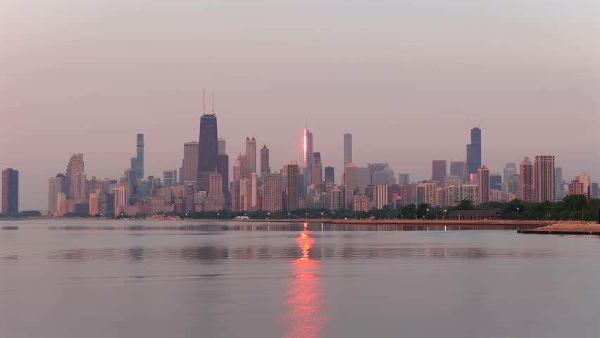 Smooth aerial rise over Lake Michigan, holding the hazy Chicago skyline centered as a bright sunrise flare and shimmering reflection gradually fade across the calm water during a smoky summer morning