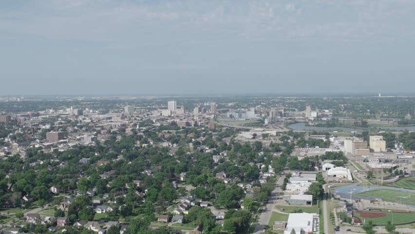 Aerial flyover glides toward Cedar Rapids Iowa downtown skyline showing city buildings, river and residential neighborhood with dense tree canopy