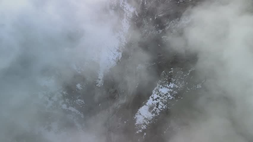 Top down drone view of fog drifting through a snowy mountain canyon with a river below. Winter aerial footage of a rugged mountain landscape.