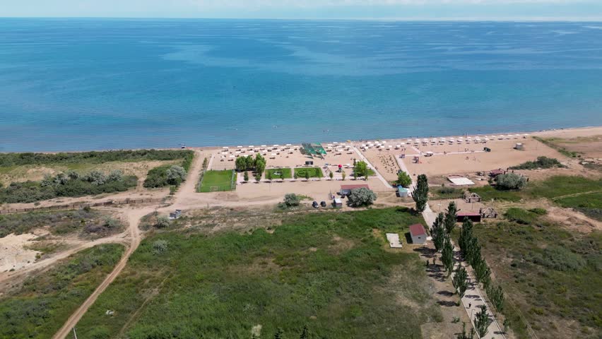 Drone view of a lakeside resort on the shore of Issyk-Kul Lake, Kyrgyzstan, with a beach, shoreline and resort buildings surrounded by natural landscape in summer.