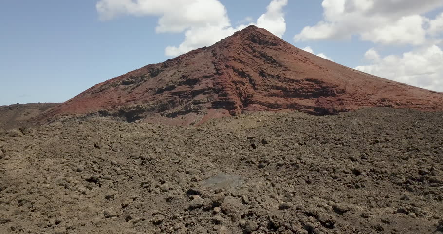 4K aerial drone footage ascending over a rugged Malpais lava field to reveal a striking red volcanic cone in the Montañas del Fuego, Timanfaya National Park, Lanzarote. The shot curves slightly before rising vertically to 100 meters, showcasing the sharp aa lava rocks, geological strata, iron oxidation, and the vast Martian-like volcanic landscape in the background.