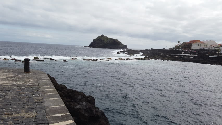 woman with long hair standing on a stone pier, looking at the rough Atlantic Ocean and the Roque de Garachico islet in Tenerife