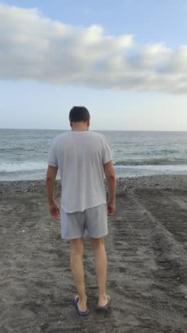 Solitary man viewed from behind walking on a dark sand beach towards the sea, stopping at the shoreline to contemplate the calm water and cloudy sky during a peaceful summer evening