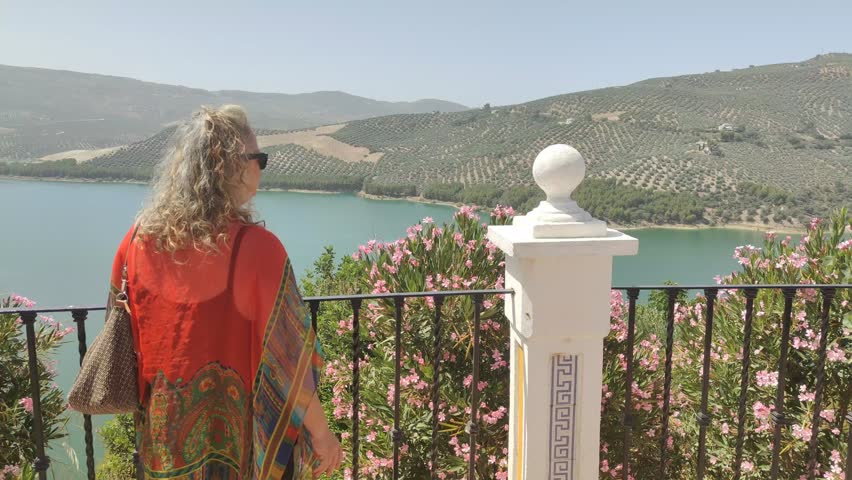 Mature woman with grey hair admiring a stunning view of a serene lake and mountains covered with olive groves from a sunny terrace with blooming pink oleanders, enjoying her vacation