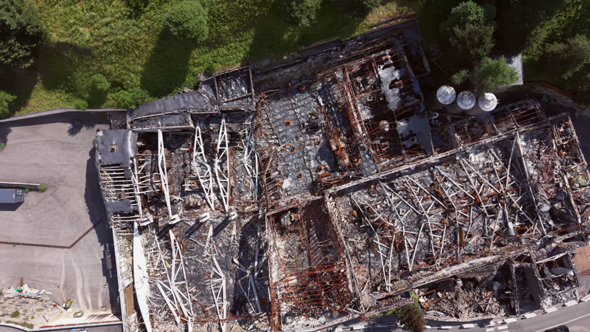 Top-down view of a building completely destroyed by fire, revealing charred structural steel beams, ash-covered debris, collapsed roof and burned-out interior ruins