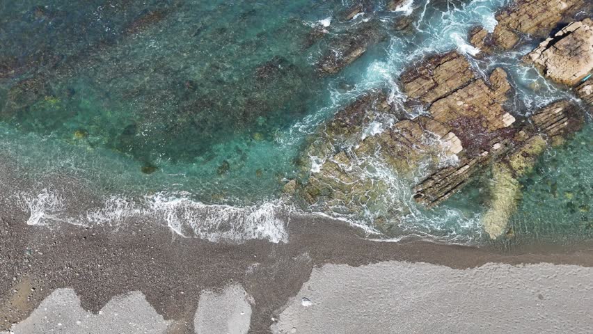Aerial top down view of turquoise ocean waves washing over rocks and pebble beach