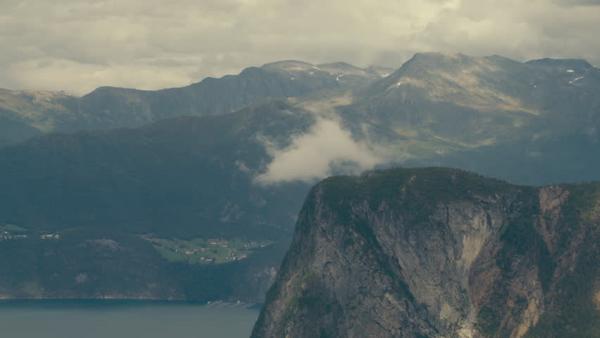 Steep cliff rises above a calm Norwegian fjord as low clouds drift across layered Sunnmøre mountains.