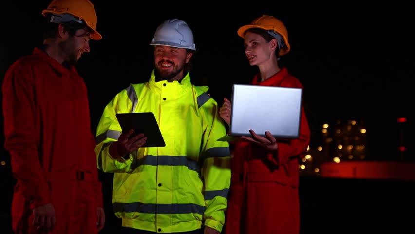 Teams engineers gas separation plants petrochemical plants stand together meetings during night shift holding tablets check data plan engineering work amidst the brightly lit modern energy industrial structure that operates 24 hours day.
