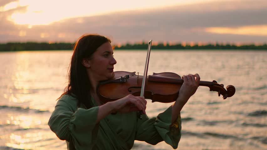 Violinist performing by tranquil lakeshore at golden sunset, serene woman playing classical music outdoors with warm evening light reflecting on water surface
