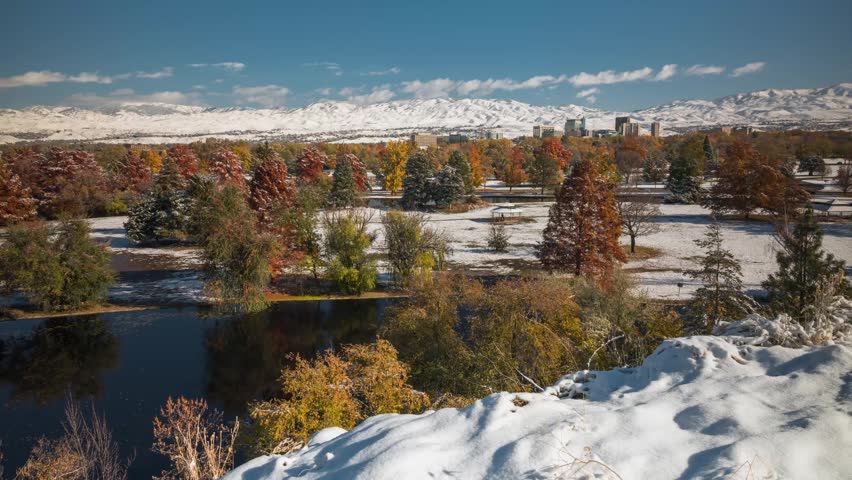 Aerial view of Ann Morrison Park featuring snow-covered trees, a pond, and the Boise city skyline against a backdrop of snow-capped mountains in Boise, Idaho, United States.
