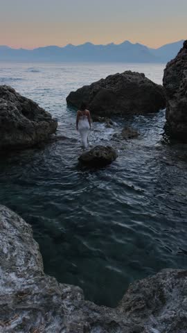 Cinematic mysterious woman in white dress standing among coastal rocks at blue hour with calm sea atmospheric surreal emotional scene, vertical