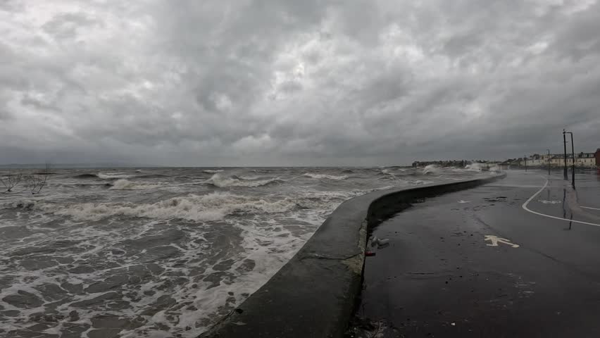 Stormy waves crash along a wet coastal road under heavy gray clouds, reflecting the dramatic seaside weather as Storm Bram hits Scotland