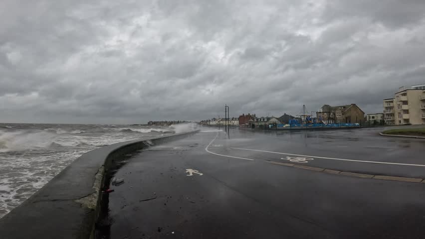 Stormy waves crash along a wet coastal road under heavy gray clouds, reflecting the dramatic seaside weather as Storm Bram hits Scotland