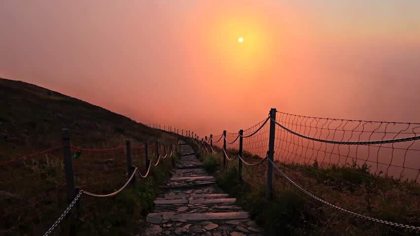 Horizontal view of a stone paved mountain trail with chain safety fence enveloped in thick pink and orange sunset fog and low clouds on Sniezka.