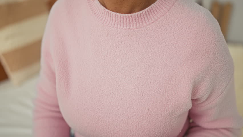 Woman with hands on chest touching pink sweater while seated on bed in bedroom; comfort gratitude healing.