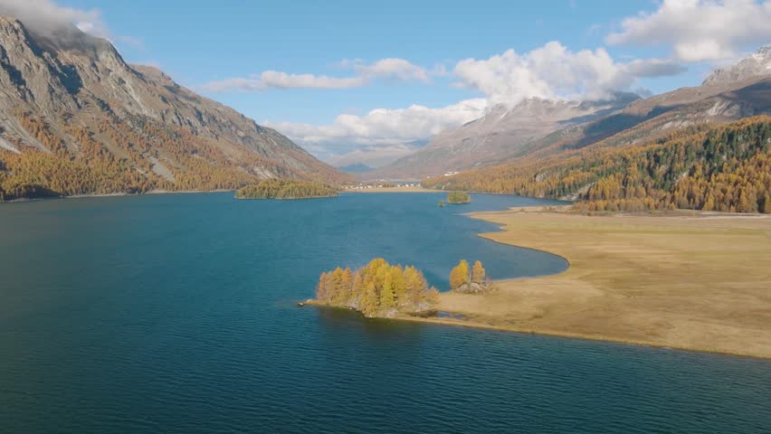 Aerial drone video of mountains and a lake in Engadin valley, Switzerland. Sunny autumn landscape with golden trees and water reflection in motion.