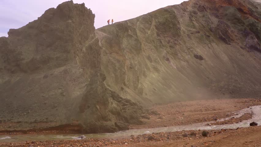 Aerial view capturing two hikers walking along sharp volcanic mountain ridge overlooking fast flowing glacial river in Icelandic highlands. Dynamic cinematic footage showing rugged terrain.