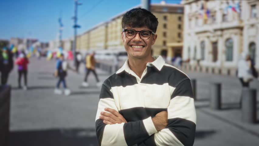 Young hispanic man wearing glasses with crossed arms standing on a sunlit street; easygoing confidence.