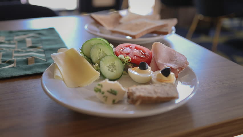 Close up view of a fresh and healthy breakfast plate served on a wooden table. The platter includes sliced ham, cheese, crisp cucumber slices, a cherry tomato, and toasted bread, capturing a cozy