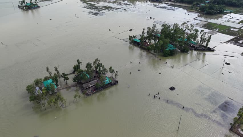 Aerial view of flooded rural landscape with houses on elevated ground and people wading through water in Banshkhali Upazila, Chittagong Division, Bangladesh.