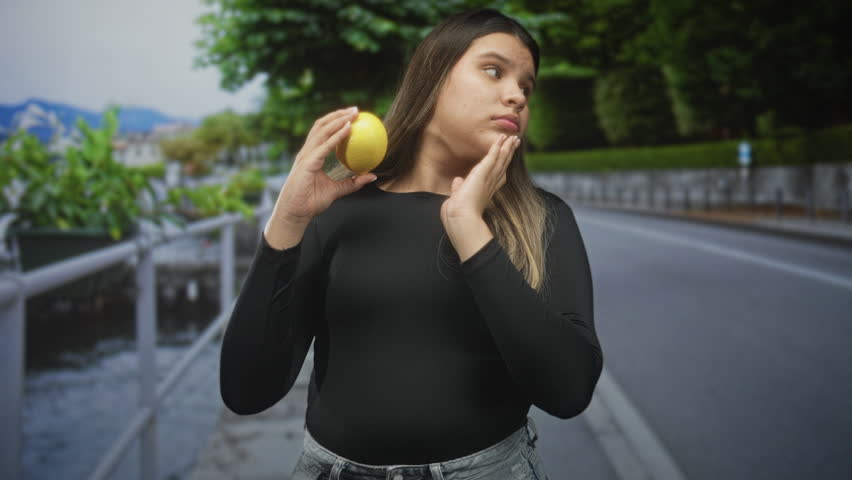 Young woman holds lemon near mouth with fingers, pensive look on street by canal walkway; wellness curiosity.