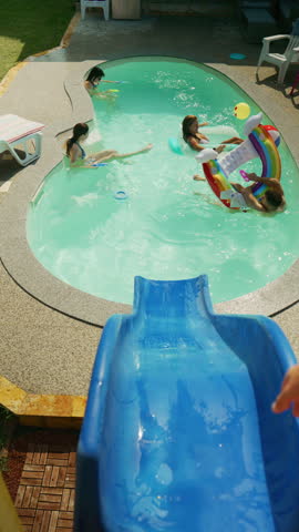 A man slides down a blue water slide into a backyard swimming pool. People enjoy swimming and relaxing on inflatable floats on a sunny day.