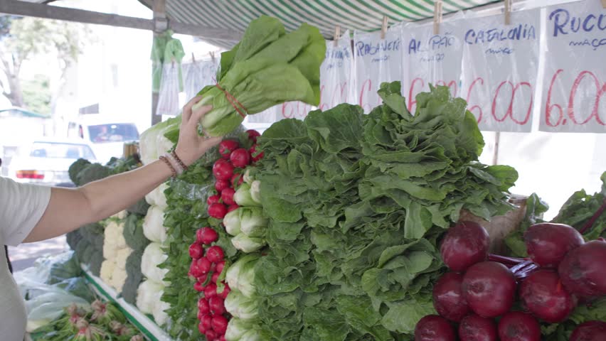 A woman buys fresh organic vegetables and greens at an open-air market stall. Open-air food market; a traditional street market in São Paulo.