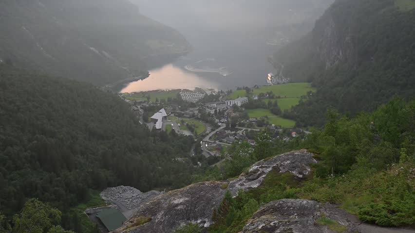 Elevated view of Geirangerfjord, Norway, with green fields, small town, winding roads, and calm water between steep forested mountains.