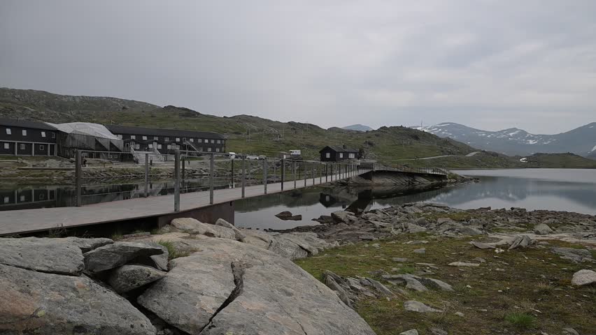 Rocky alpine plateau with still mountain lake and distant snowcapped peaks under overcast summer sky in Norway.