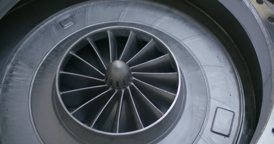 A close view shows a large metallic running wheel with numerous curved spokes turning steadily around a central hub. The silvery wheel sits inside a round housing and forms the heart of an industrial machine in a steel factory.