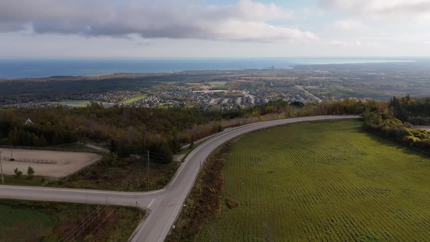 A drone view of a white car rounding a hilltop bend above Collingwood, Ontario, Canada overlooking a lakeside village and autumn landscape