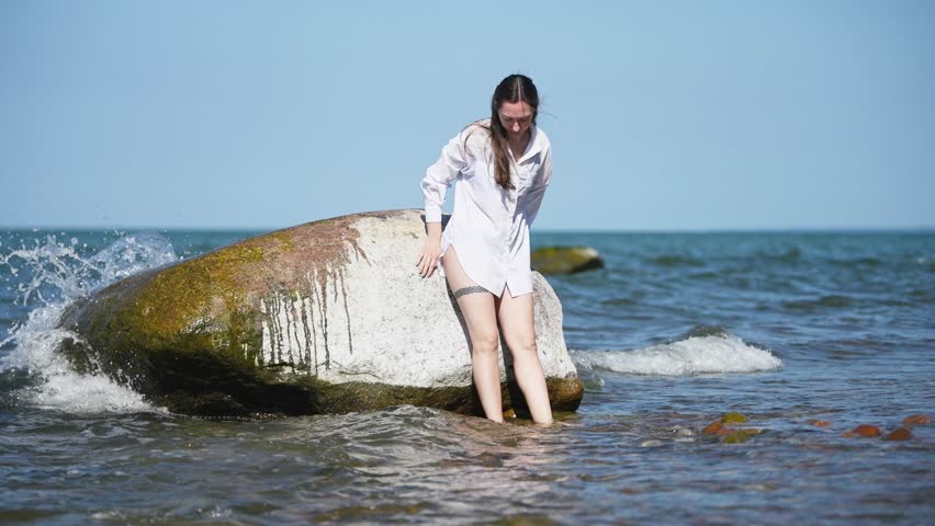 Woman in white shirt stands and poses next to mossy sea rock in calm shallow ocean waters with gentle waves and clear blue sky background during sunny day