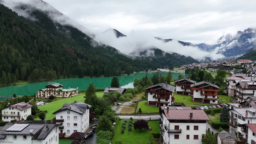 Aerial drone shot flying over turquoise alpine lake towards mountain valley village with clouds touching green forest peaks on overcast day