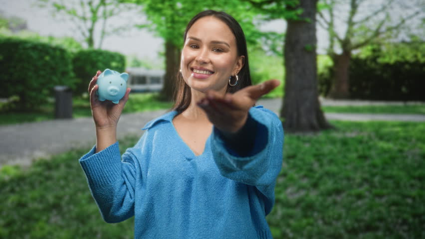 Woman holding a blue piggy bank in her left hand and extends open right hand toward camera on street setting, smiling; savings optimism.