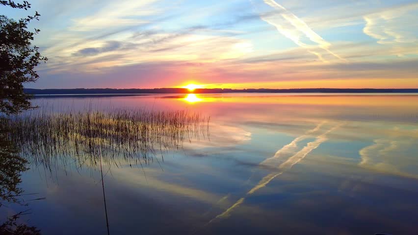 Clouds move across the sky as the sun sets over the lake. The water reflects the colors of the sky, and grasses show along the shore. This scene captures the end of the day.