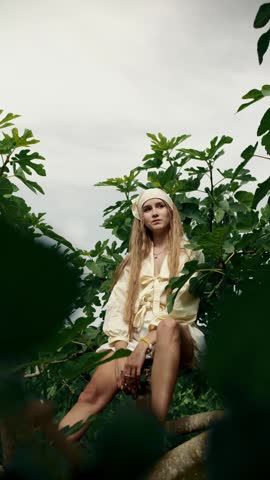 Beautiful young woman sitting on a tree branch, surrounded by green leaves. She opens a ripe fig and enjoys eating the fresh fruit