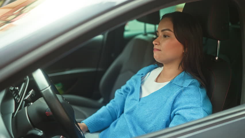 Young hispanic woman clutching steering wheel with head bowed on the wheel, wearing blue sweater in a parked car on a street; frustration recovery.