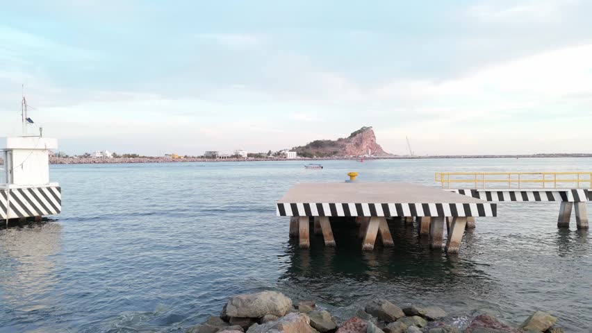 View of the entrance to Mazatlan Harbor from the pier, featuring a small fishing boat crossing calm waters toward Stone Island (Isla de la Piedra), with Cerro de los Chivos in the background. Peaceful coastal landscape under a soft blue sky in Sinaloa, Mexico.