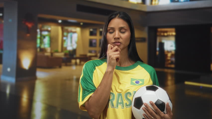 Woman in brazil soccer jersey holding a soccer ball and making a palm up questioning gesture inside a building lobby; national pride contemplation.