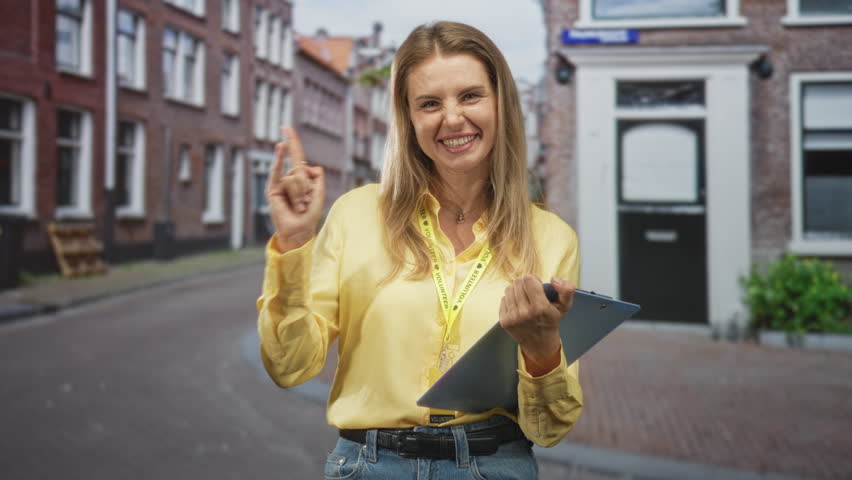 Woman holding clipboard and visitor card, raises index finger and displays hand while smiling in a narrow street with building doorway and pavement; friendly welcome.