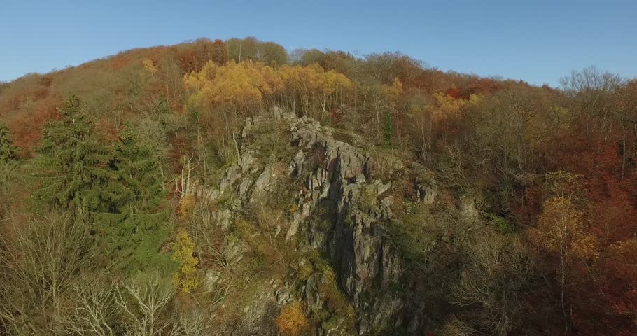 A rugged rock cliff rises out of a dense forest that glows in vivid orange and golden autumn colours beneath a clear blue sky.
