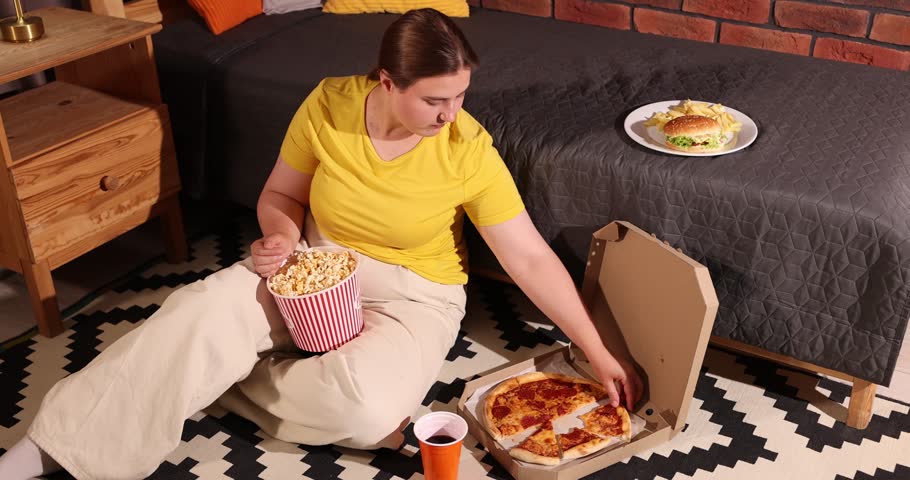 Overweight woman with bucket of popcorn eating pizza on floor near bed at home