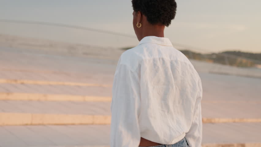 Black African American woman walks along the waterfront promenade during golden hour with the sea in the background.