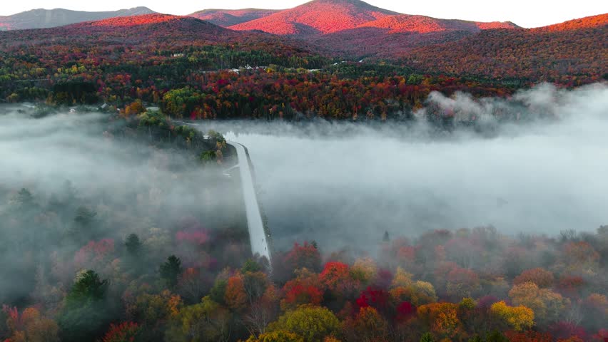 A drone view of colorful mountain forests in New England as early morning fog drifts through valleys and autumn hills