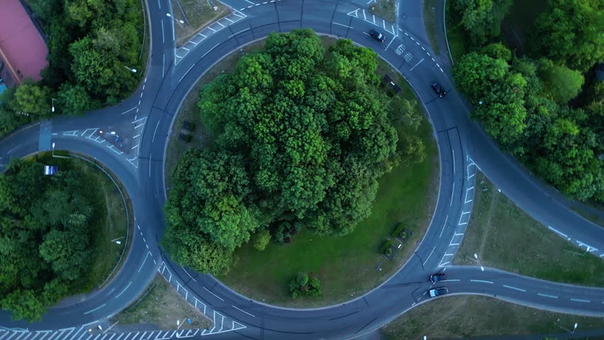 Top down aerial perspective of roundabout intersection with cars driving through circular road network amidst green lush trees in urban environment during twilight time motion footage.