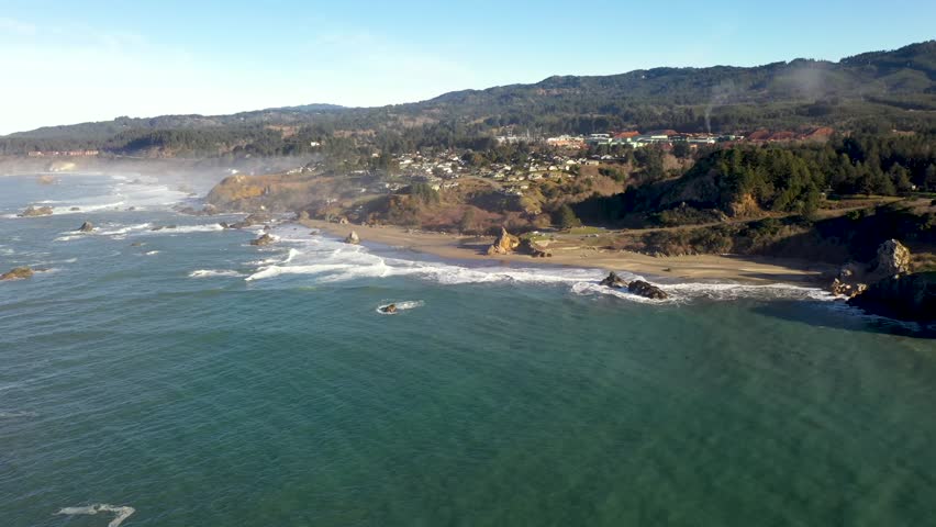 Aerial drone cinematography captures panoramic landscape of rugged Oregon coastline with turquoise water waves rolling onto sandy beach near coastal town and evergreen forest hills.