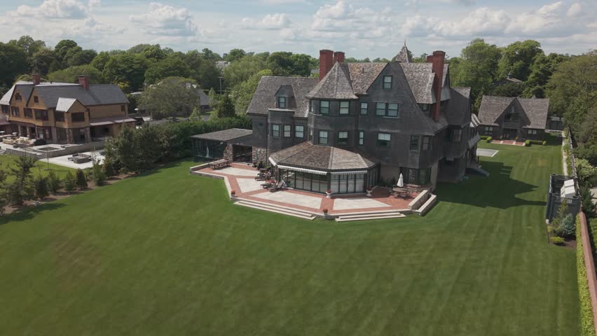 Shingle-style Victorian mansion features turret plus chimneys above octagonal brick patio, manicured lawn stretching toward neighboring estate in Newport, Rhode Island. Aerial drone view.